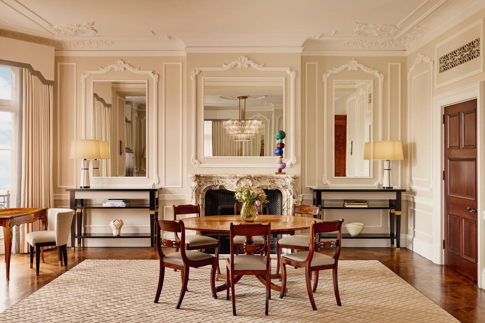 Refined dining room featuring a round wooden table with six chairs, ornate wall panelling, and a marble fireplace framed by two large mirrors. Matching console tables with lamps and decorative objects stand on either side.