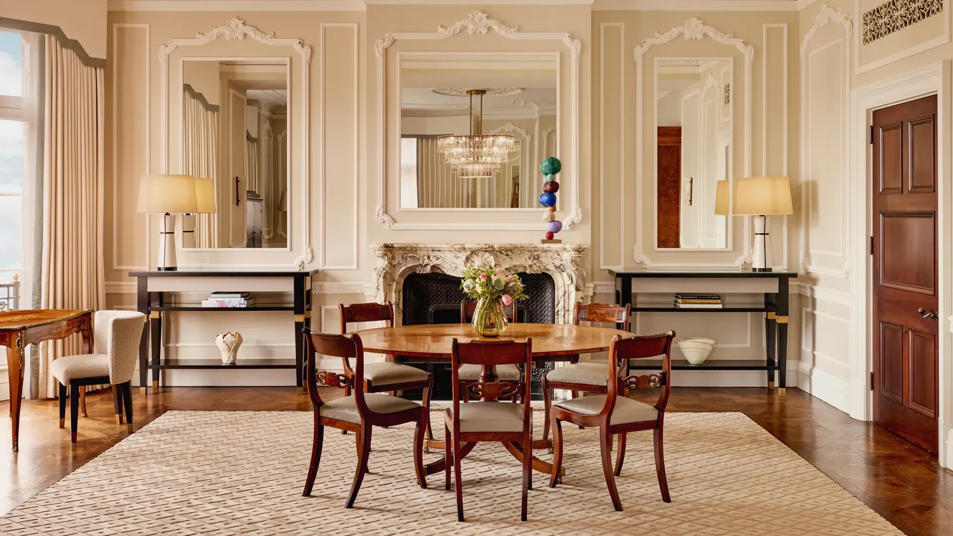 Refined dining room featuring a round wooden table with six chairs, ornate wall panelling, and a marble fireplace framed by two large mirrors. Matching console tables with lamps and decorative objects stand on either side.