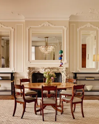 Refined dining room featuring a round wooden table with six chairs, ornate wall panelling, and a marble fireplace framed by two large mirrors. Matching console tables with lamps and decorative objects stand on either side.