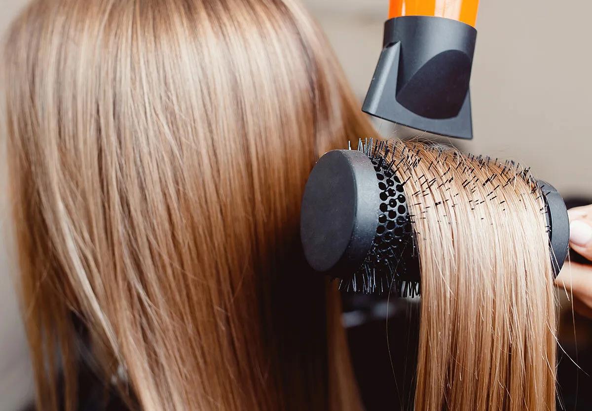 
Close-up of smooth blonde hair being styled with a round brush and blow dryer during a professional salon blowout treatment.
