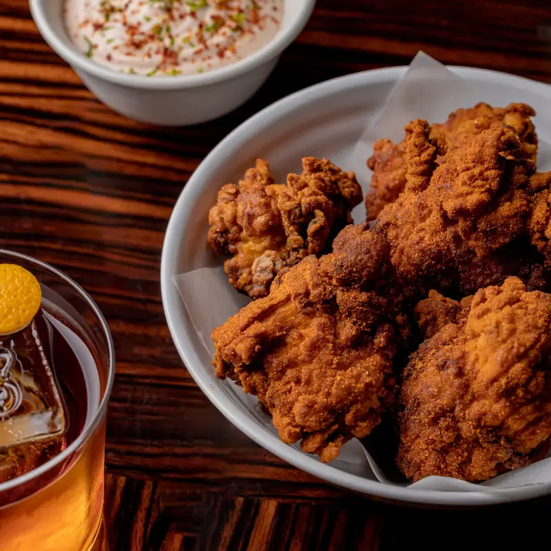 Close-up of a bowl of crispy fried chicken pieces served with a side of creamy dipping sauce topped with herbs, and a glass of amber cocktail with a citrus garnish on a polished wooden table.