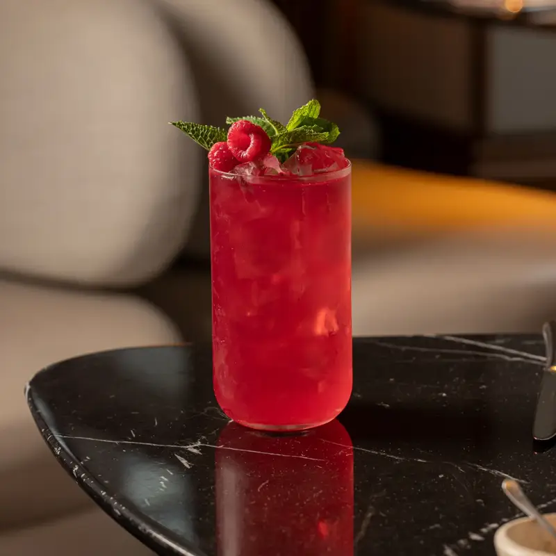 Bright pink cocktail garnished with raspberries and mint on a dark marble table.