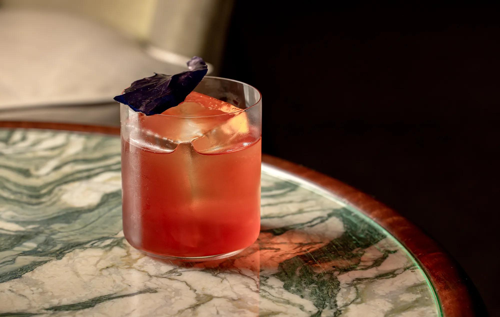 Pink cocktail with large ice cube and purple leaf garnish on green marble table.