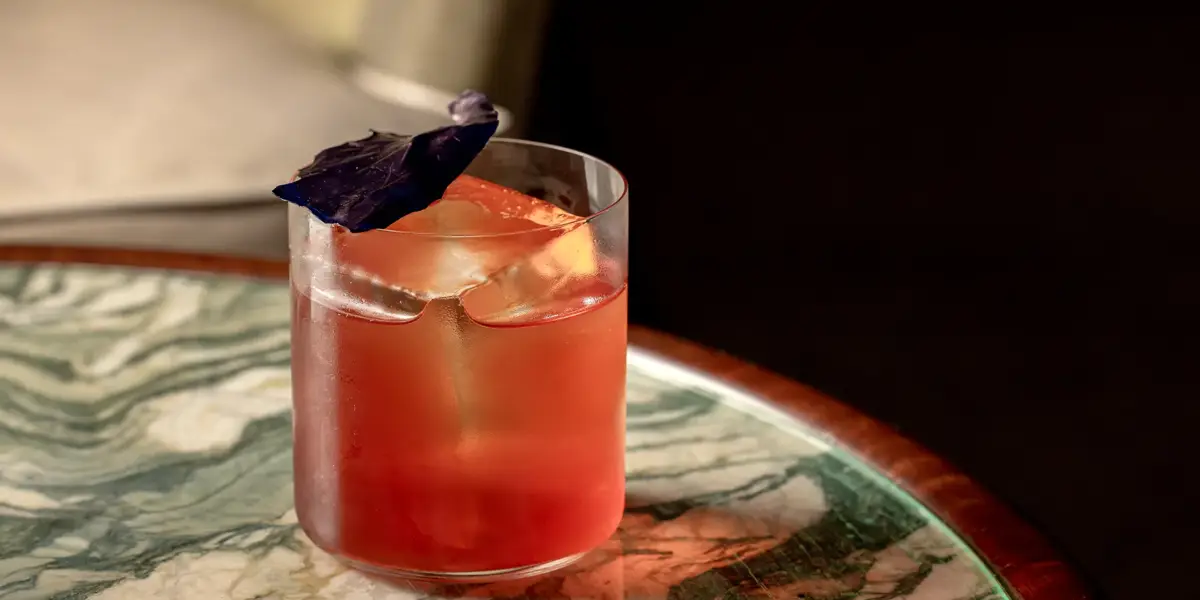 Pink cocktail with large ice cube and purple leaf garnish on green marble table.