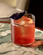 Pink cocktail with large ice cube and purple leaf garnish on green marble table.