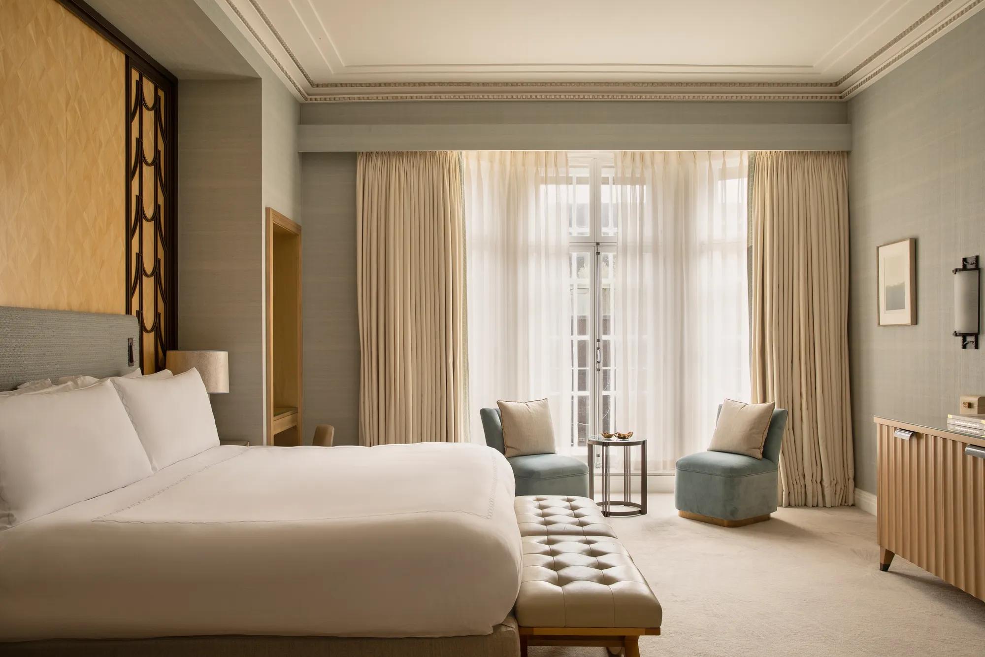 Contemporary bedroom with a king-size bed and upholstered bench, facing two blue armchairs and a side table beside floor-to-ceiling windows with sheer curtains.