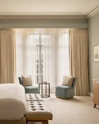 Contemporary bedroom with a king-size bed and upholstered bench, facing two blue armchairs and a side table beside floor-to-ceiling windows with sheer curtains.