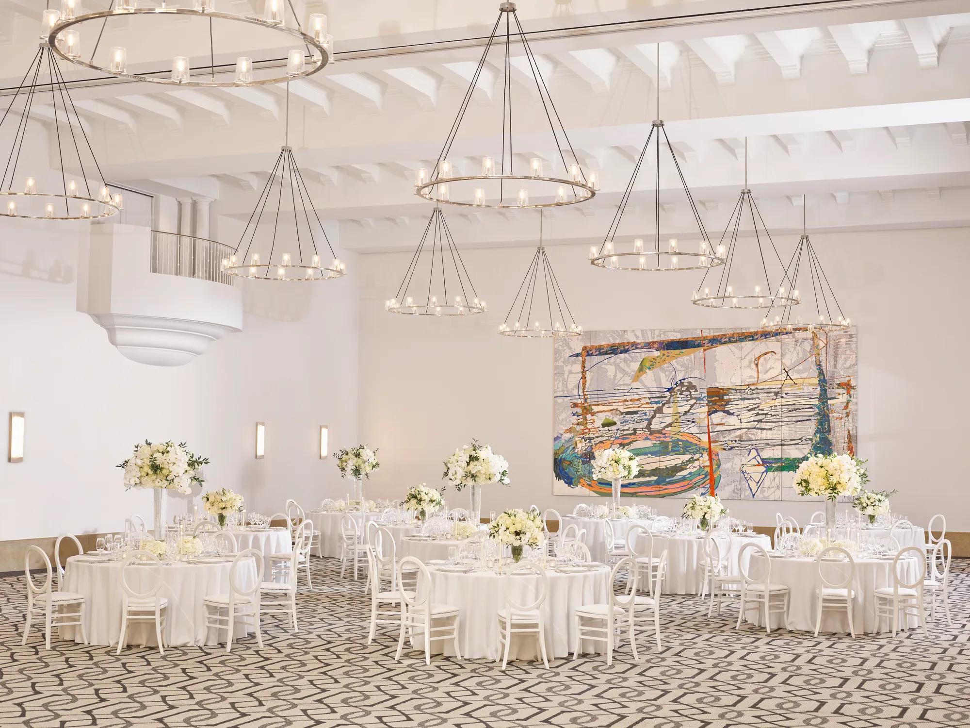 Daylit ballroom in banquet rounds; white linens and tall white florals, abstract mural and tiered ring chandeliers overhead.