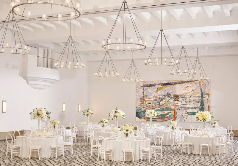 Daylit ballroom in banquet rounds; white linens and tall white florals, abstract mural and tiered ring chandeliers overhead.