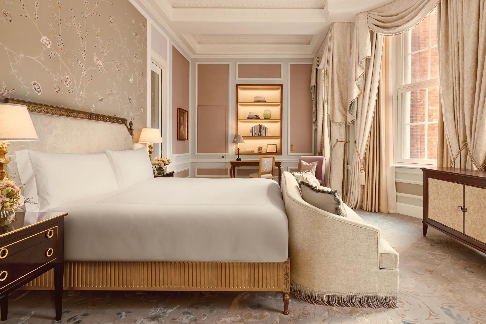 Elegant bedroom with a king-size bed, embroidered floral wallpaper, and soft beige drapery, featuring a writing desk, armchair, and large windows with views of red brick architecture outside.