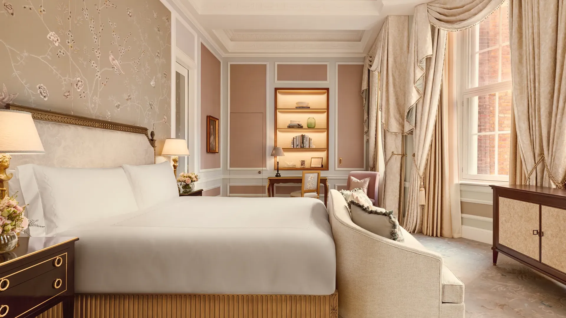 Elegant bedroom with a king-size bed, embroidered floral wallpaper, and soft beige drapery, featuring a writing desk, armchair, and large windows with views of red brick architecture outside.