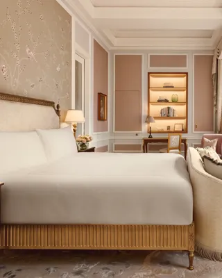 Elegant bedroom with a king-size bed, embroidered floral wallpaper, and soft beige drapery, featuring a writing desk, armchair, and large windows with views of red brick architecture outside.