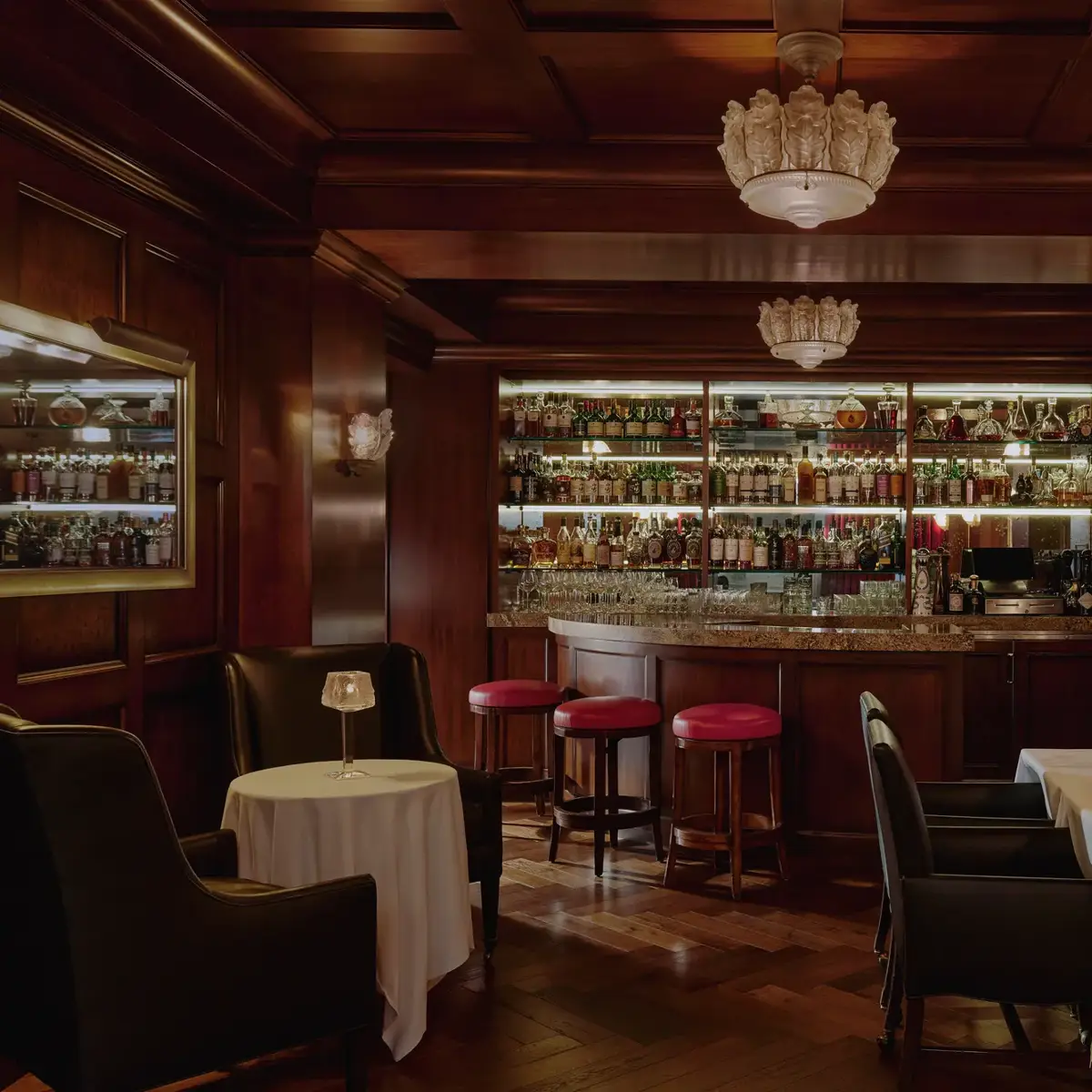 Dimly lit whisky bar with wood paneling, bottle-lined back bar, red stools, and leather seating.