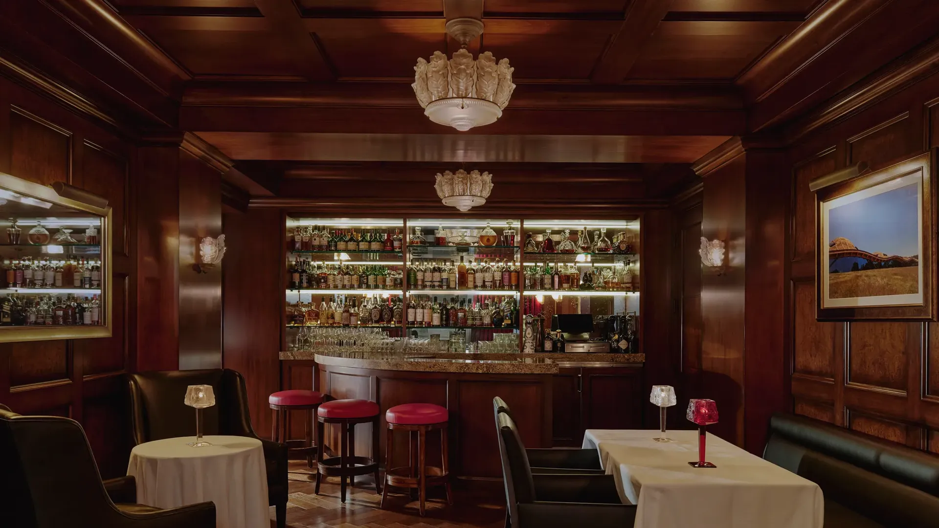 Dimly lit whisky bar with wood paneling, bottle-lined back bar, red stools, and leather seating.