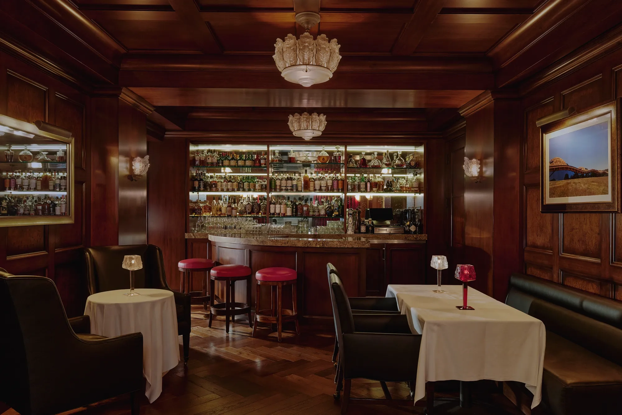 Dimly lit whisky bar with wood paneling, bottle-lined back bar, red stools, and leather seating.