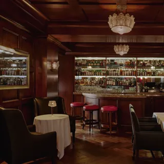 Dimly lit whisky bar with wood paneling, bottle-lined back bar, red stools, and leather seating.