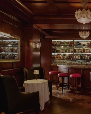 Dimly lit whisky bar with wood paneling, bottle-lined back bar, red stools, and leather seating.