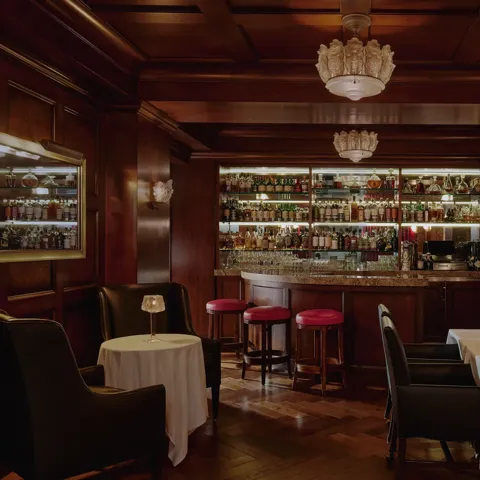 Dimly lit whisky bar with wood paneling, bottle-lined back bar, red stools, and leather seating.
