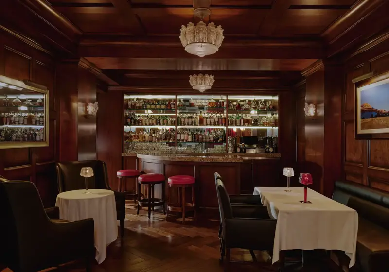 Dimly lit whisky bar with wood paneling, bottle-lined back bar, red stools, and leather seating.