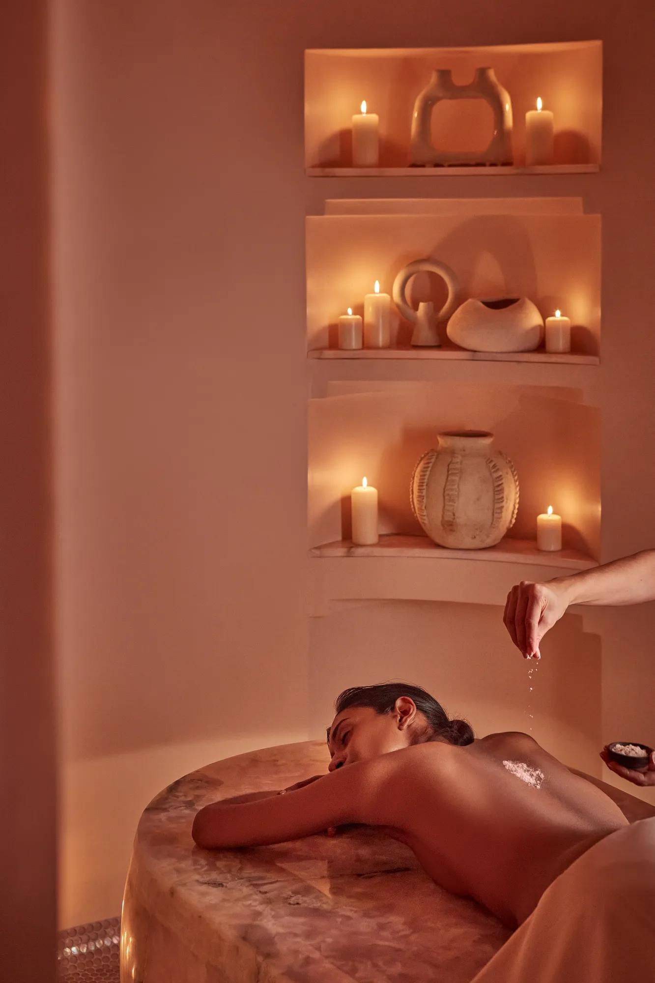Guest receiving a spa treatment on a heated stone table, surrounded by candlelit niches and sculptural ceramics.