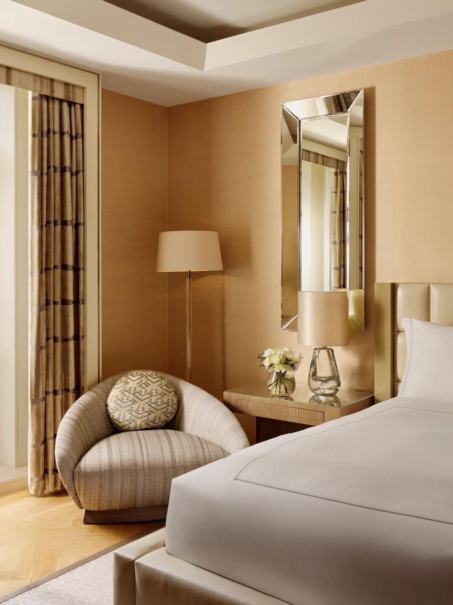 Neutral-toned bedroom corner featuring a cream upholstered bed, round striped armchair with patterned cushion, bedside table with lamp and flowers, and a tall mirror on the wall.