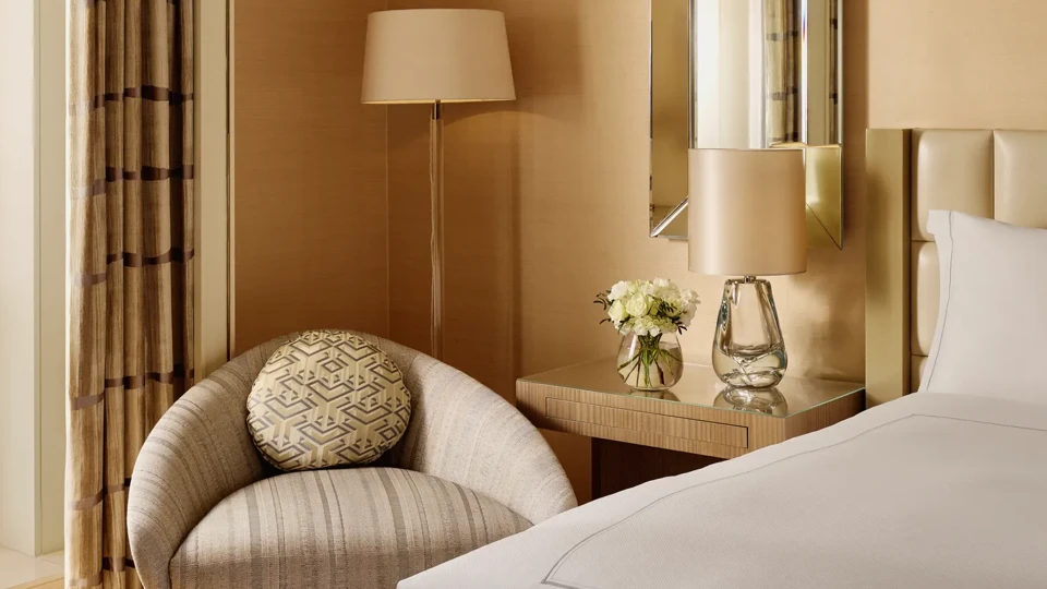 Neutral-toned bedroom corner featuring a cream upholstered bed, round striped armchair with patterned cushion, bedside table with lamp and flowers, and a tall mirror on the wall.