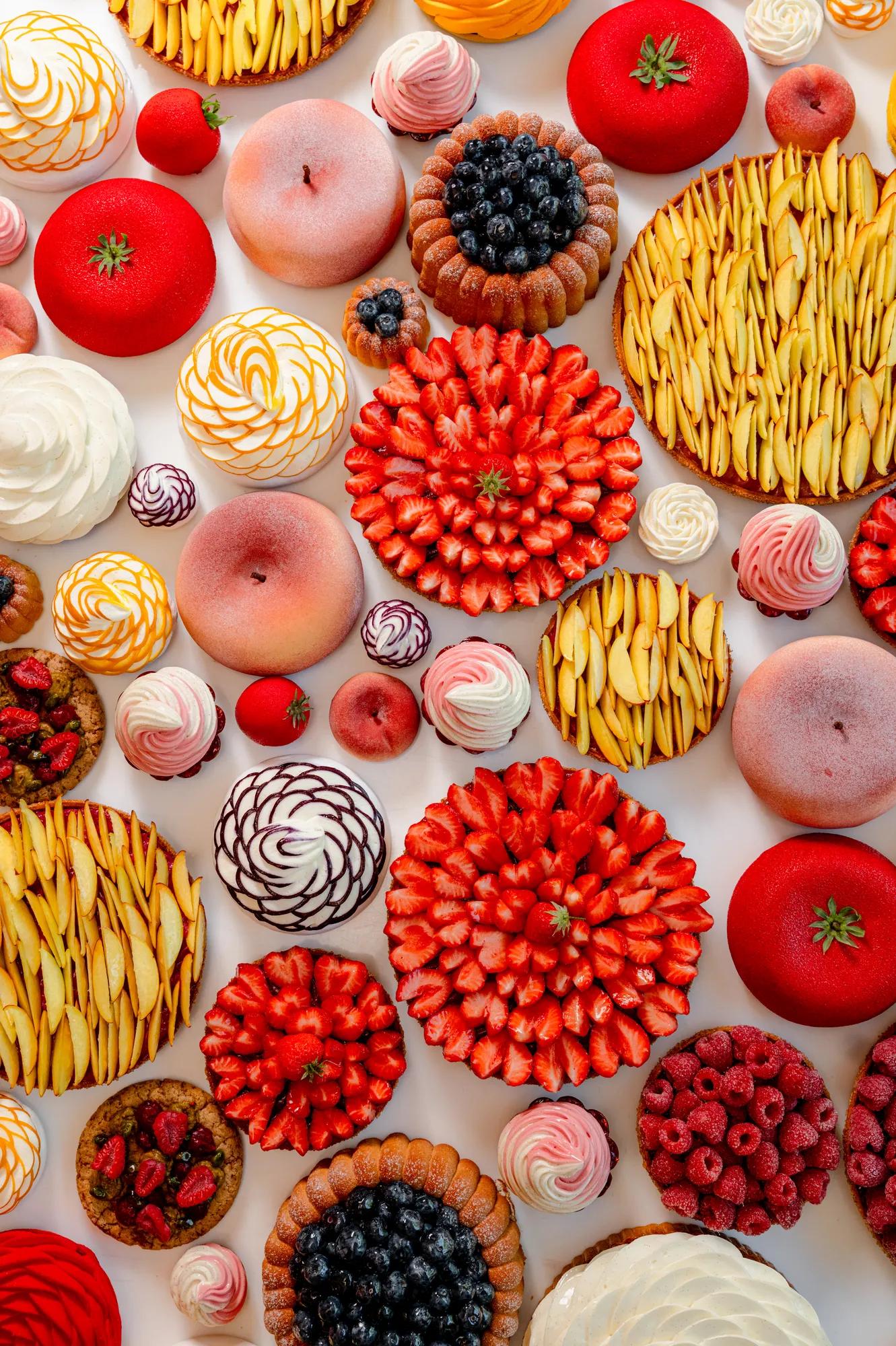 Overhead arrangement of fruit tarts, meringues and pastries featuring strawberries, peaches, blueberries and raspberries on a light background.