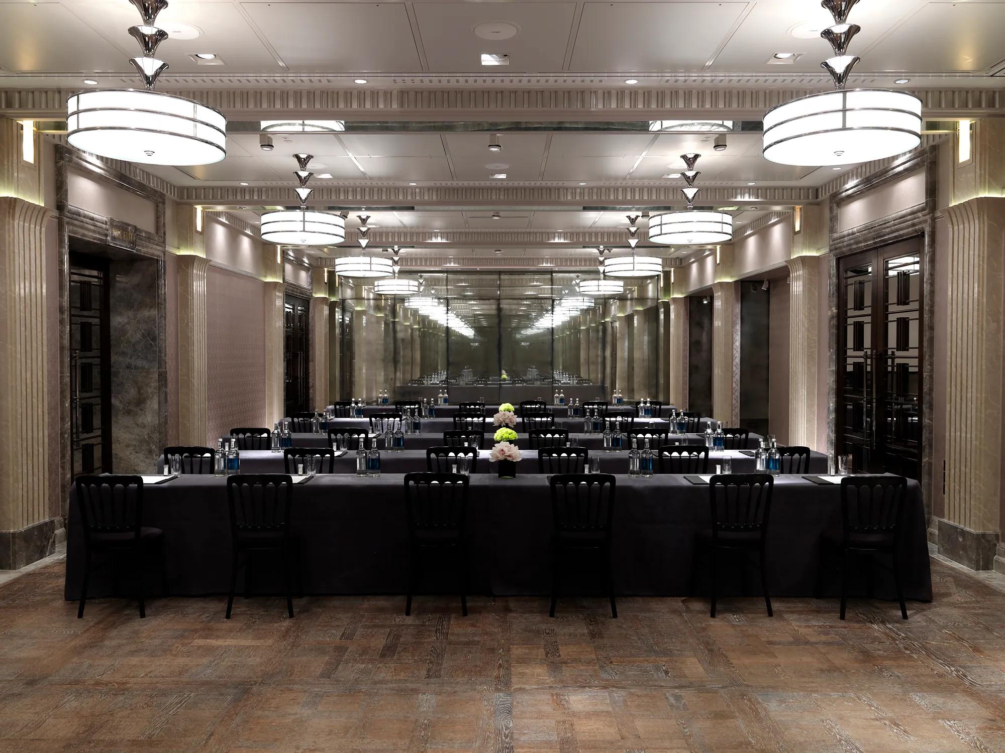 Conference room arranged in a U-shape with black-covered tables, bottled water, notepads and flowers, under mirrored walls and large Art Deco ceiling lights.