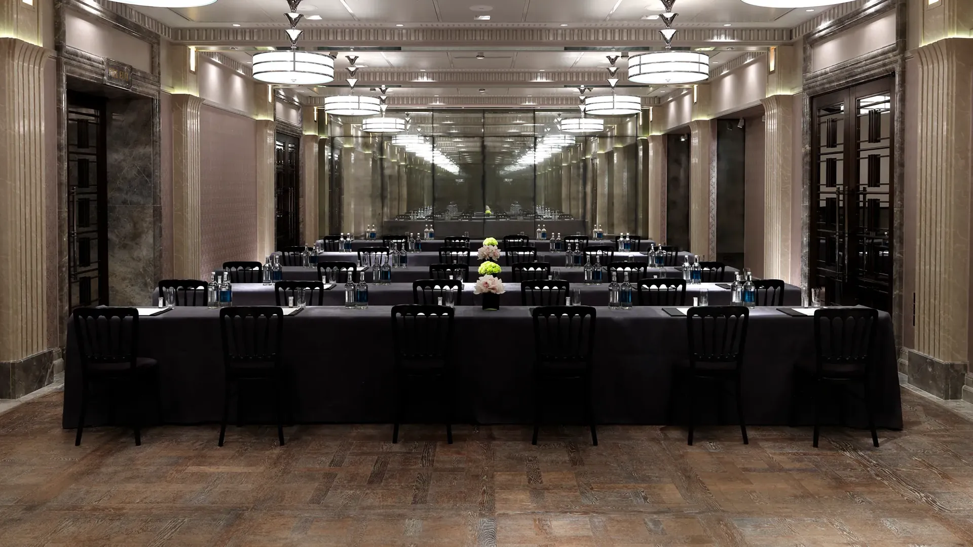 Conference room arranged in a U-shape with black-covered tables, bottled water, notepads and flowers, under mirrored walls and large Art Deco ceiling lights.