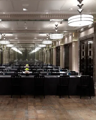 Conference room arranged in a U-shape with black-covered tables, bottled water, notepads and flowers, under mirrored walls and large Art Deco ceiling lights.