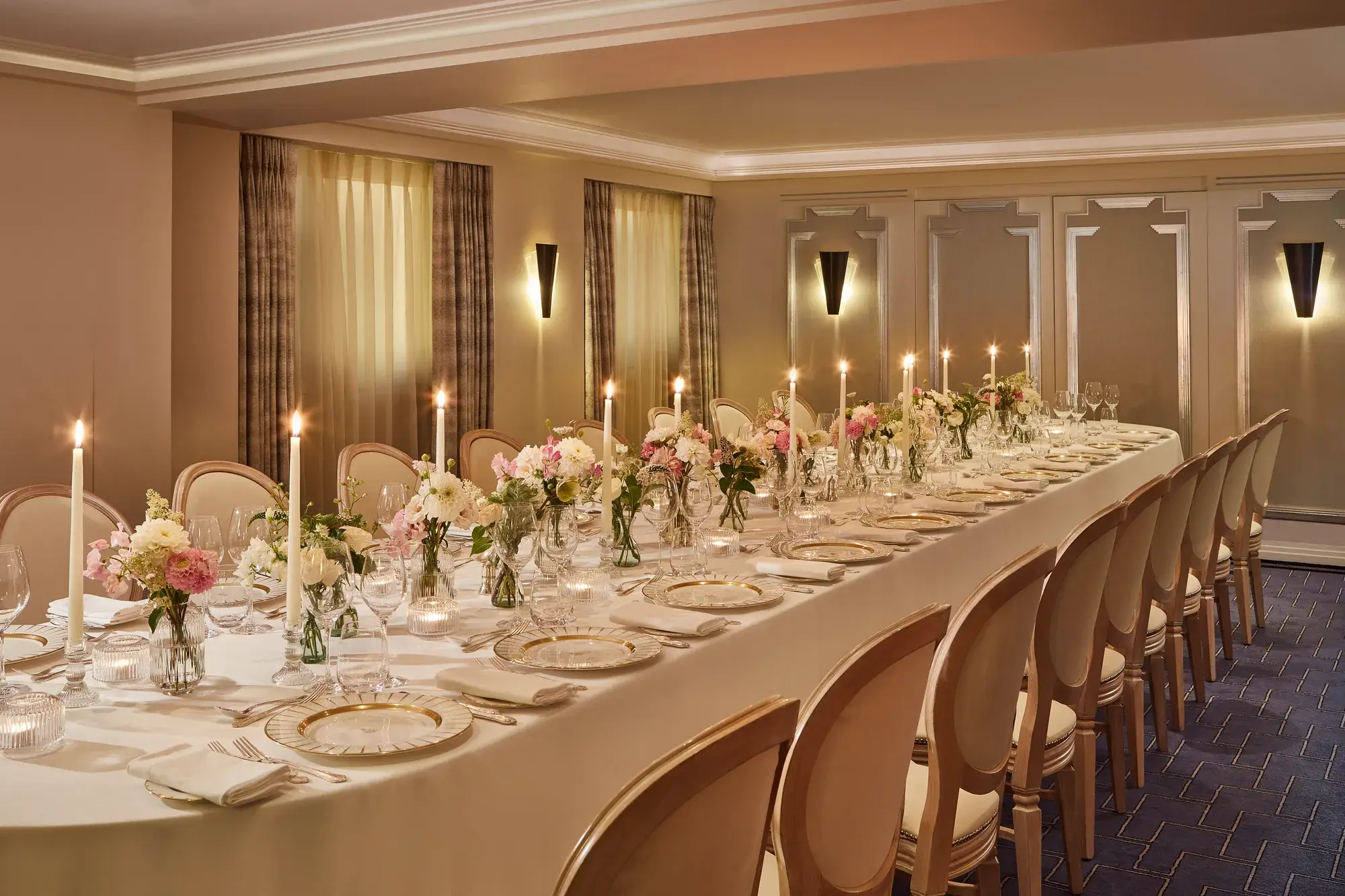 Elegant private dining room at Claridge’s with a long candlelit table set with fine china, glassware, and floral centrepieces in soft pink and white tones against taupe panelled walls.