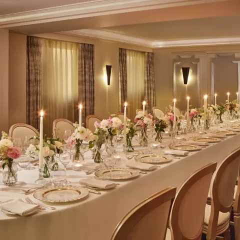 Elegant private dining room at Claridge’s with a long candlelit table set with fine china, glassware, and floral centrepieces in soft pink and white tones against taupe panelled walls.