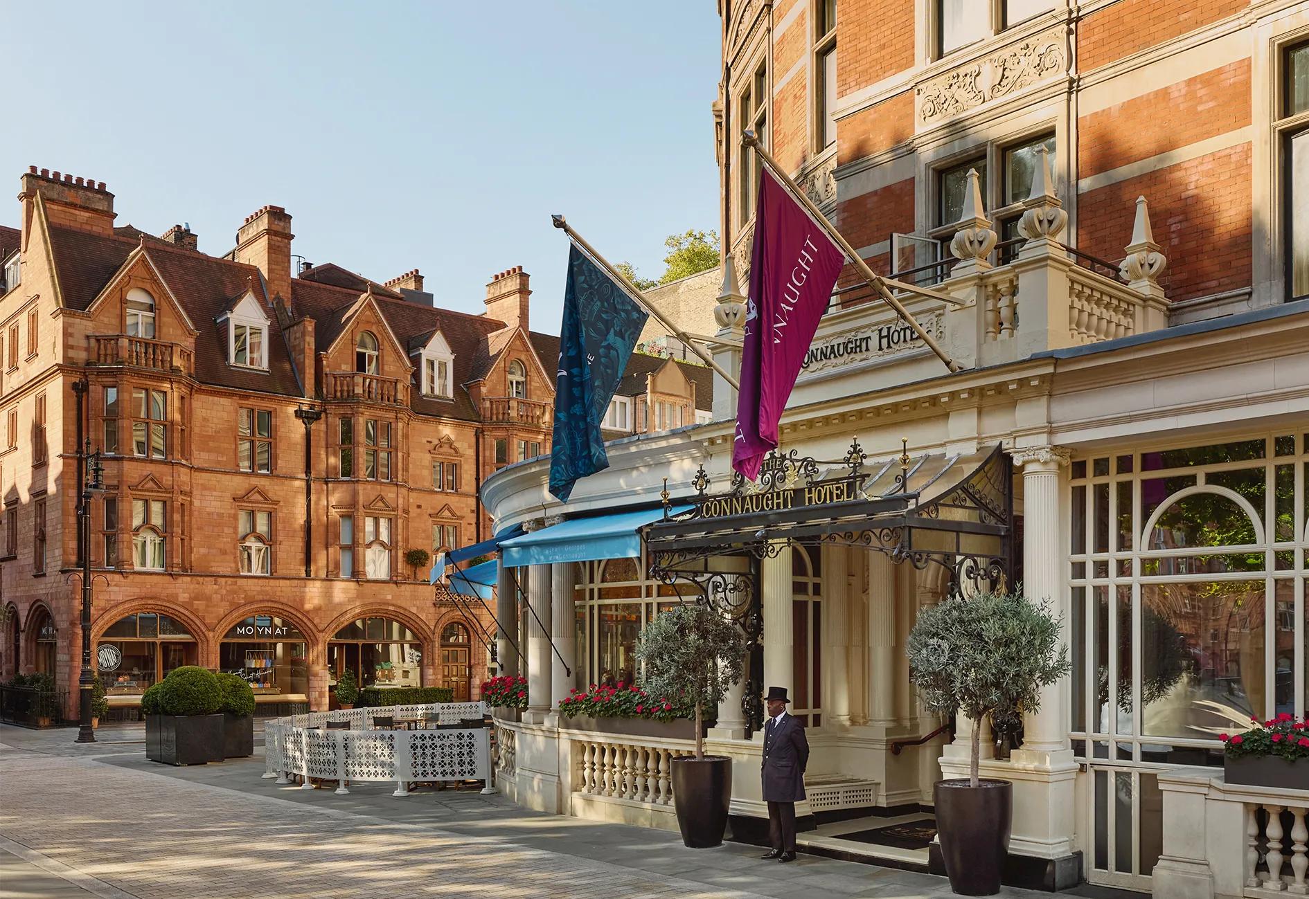 Exterior of The Connaught hotel in Mayfair, London, showing a curved red-brick façade with cream stone detailing, two flags over the entrance canopy, a doorman standing in front and potted trees by the doorway, with adjacent period buildings under a clear sky.