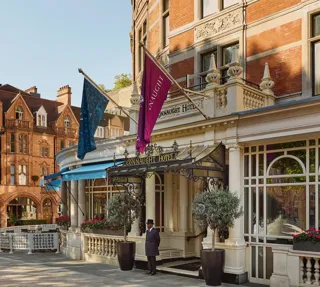 Exterior of The Connaught hotel in Mayfair, London, showing a curved red-brick façade with cream stone detailing, two flags over the entrance canopy, a doorman standing in front and potted trees by the doorway, with adjacent period buildings under a clear sky.