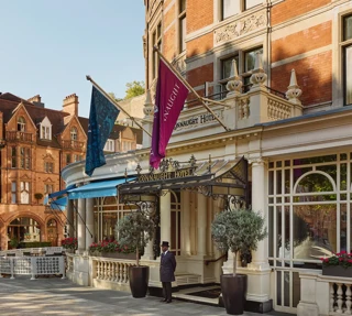 Exterior of The Connaught hotel in Mayfair, London, showing a curved red-brick façade with cream stone detailing, two flags over the entrance canopy, a doorman standing in front and potted trees by the doorway, with adjacent period buildings under a clear sky.