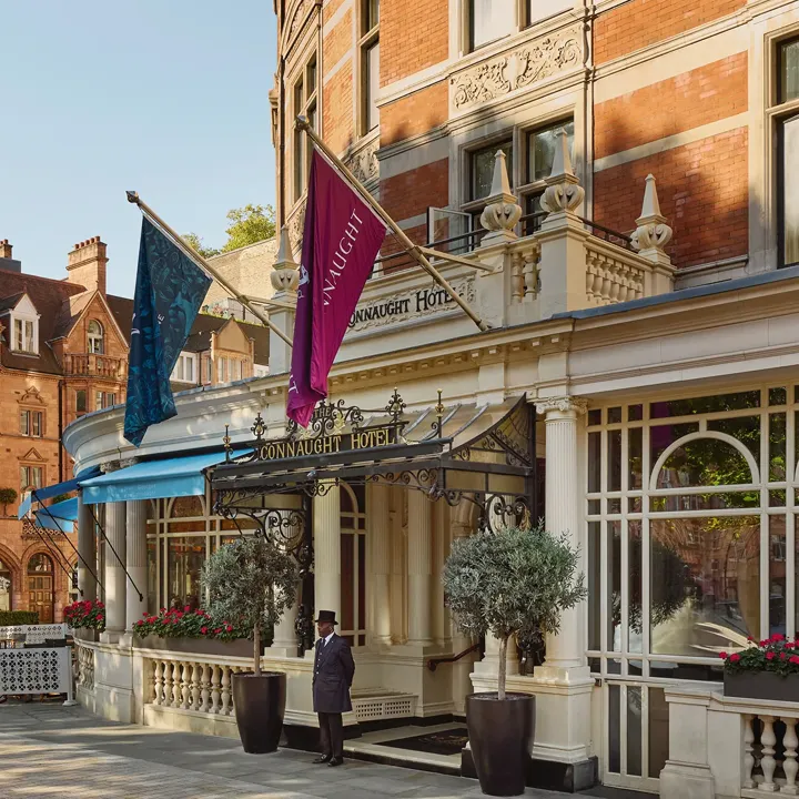 Exterior of The Connaught hotel in Mayfair, London, showing a curved red-brick façade with cream stone detailing, two flags over the entrance canopy, a doorman standing in front and potted trees by the doorway, with adjacent period buildings under a clear sky.