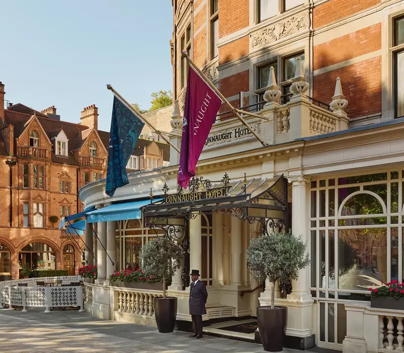 Exterior of The Connaught hotel in Mayfair, London, showing a curved red-brick façade with cream stone detailing, two flags over the entrance canopy, a doorman standing in front and potted trees by the doorway, with adjacent period buildings under a clear sky.