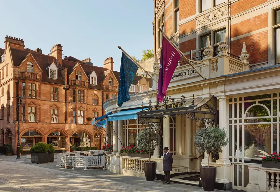 Exterior of The Connaught hotel in Mayfair, London, showing a curved red-brick façade with cream stone detailing, two flags over the entrance canopy, a doorman standing in front and potted trees by the doorway, with adjacent period buildings under a clear sky.