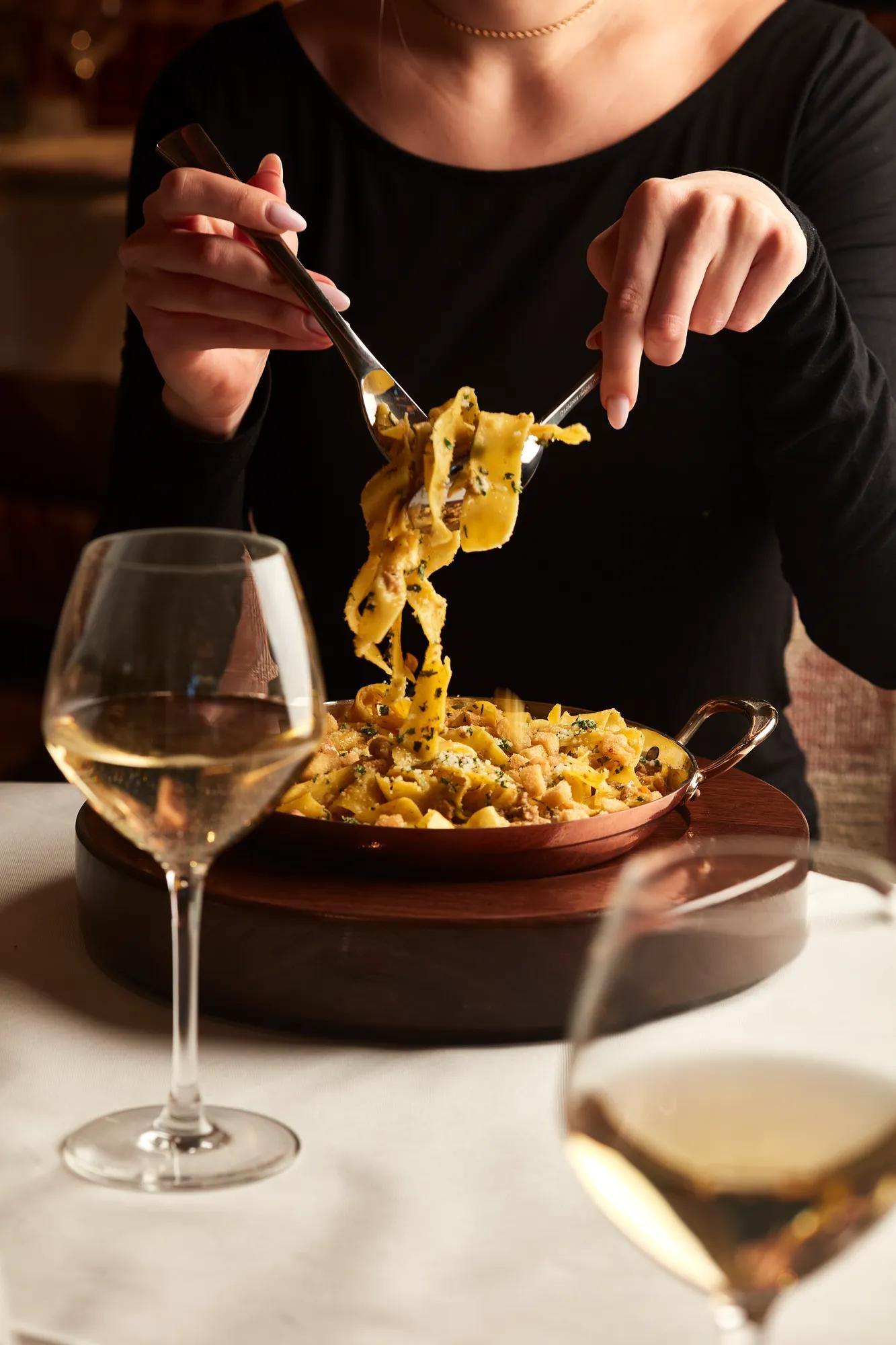 Person in black top twirling tagliatelle pasta from copper dish, with white wine glasses on table.