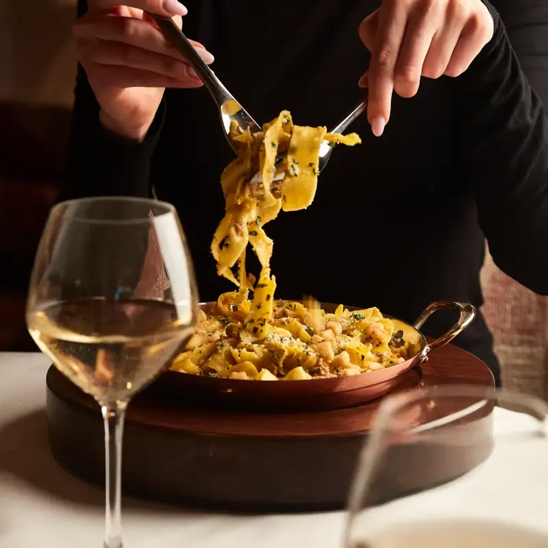 Person in black top twirling tagliatelle pasta from copper dish, with white wine glasses on table.