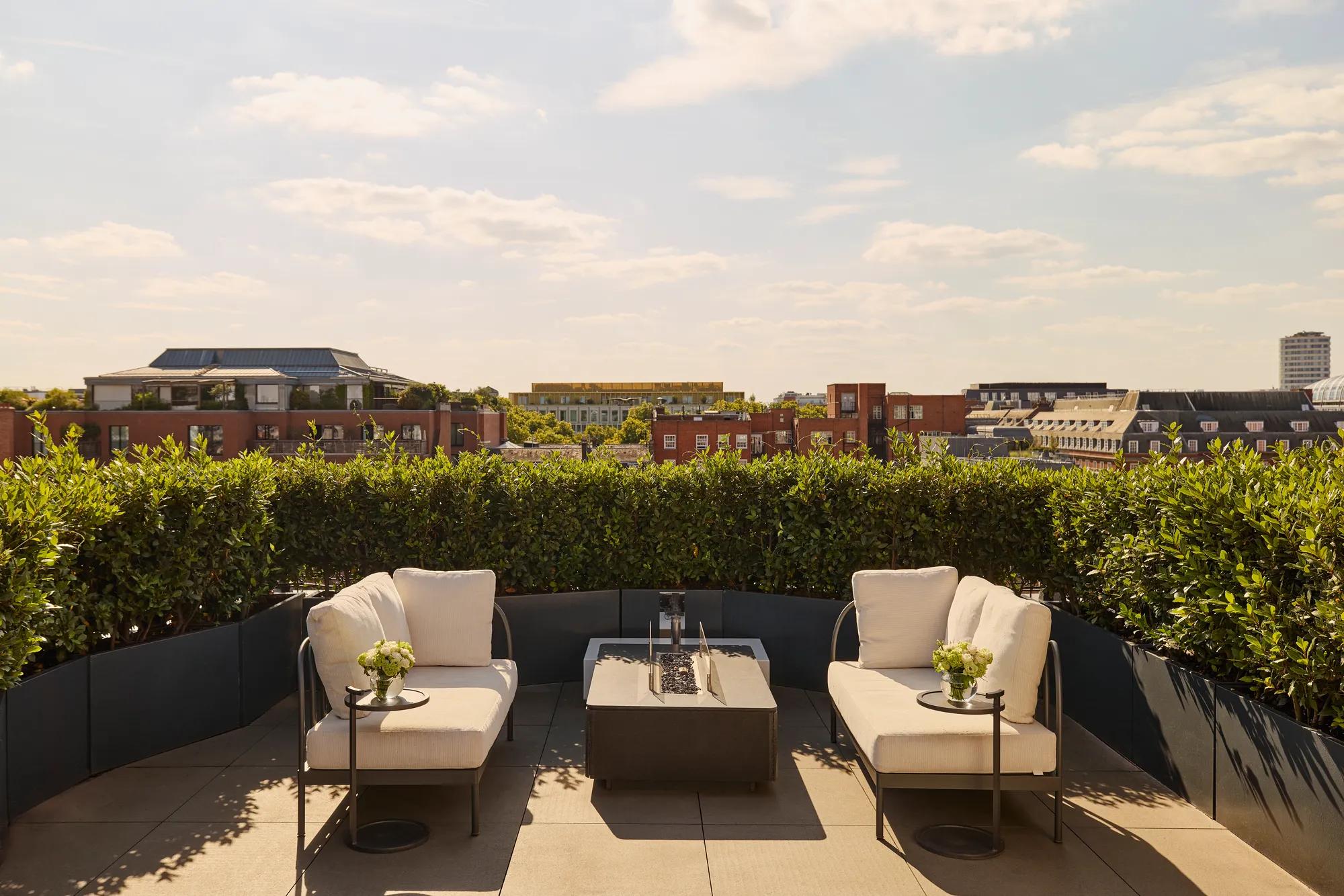 Sunny rooftop terrace with two cream lounge chairs, a central fire pit-style table, and views over red-brick buildings and greenery under a blue sky.