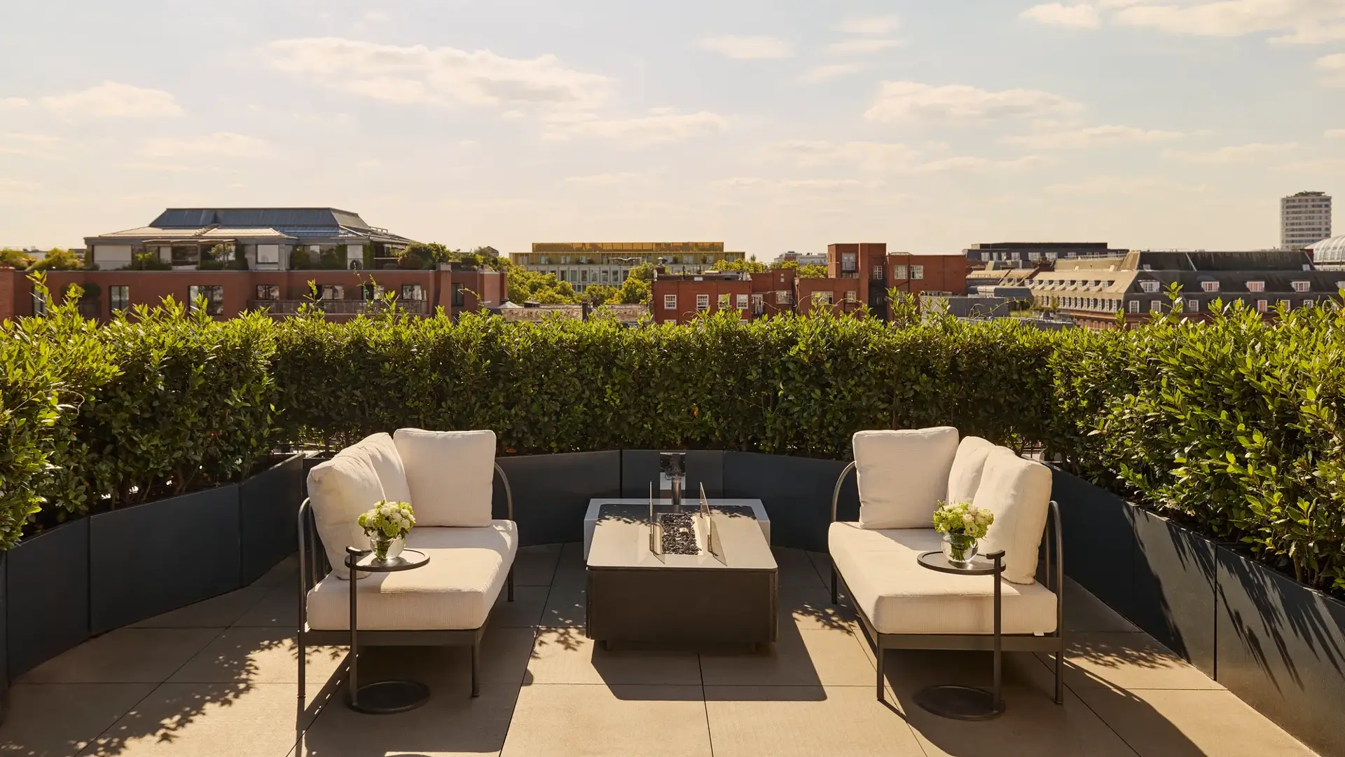 Sunny rooftop terrace with two cream lounge chairs, a central fire pit-style table, and views over red-brick buildings and greenery under a blue sky.
