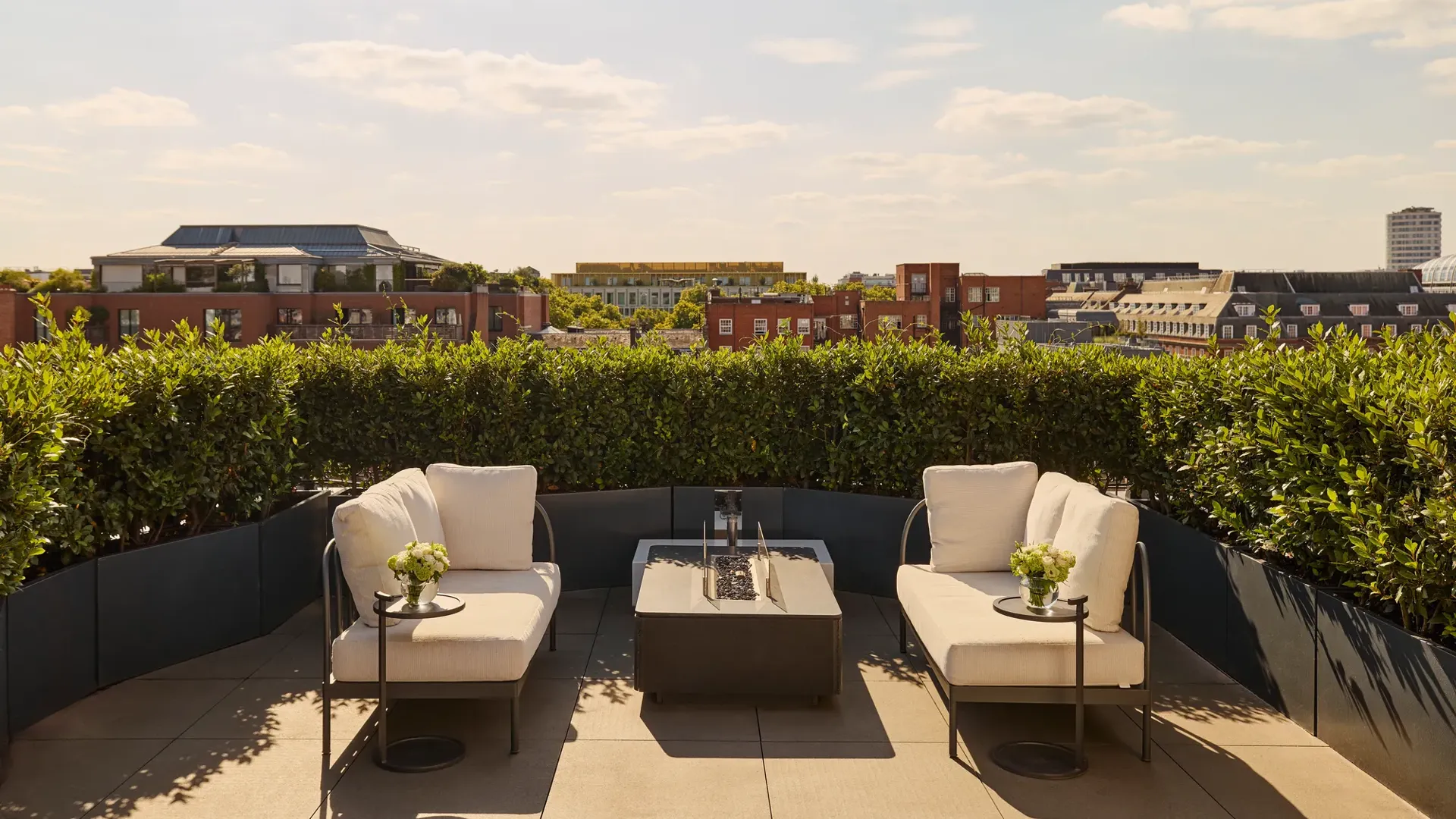 Sunny rooftop terrace with two cream lounge chairs, a central fire pit-style table, and views over red-brick buildings and greenery under a blue sky.