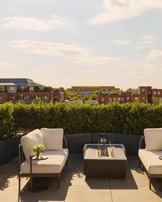 Sunny rooftop terrace with two cream lounge chairs, a central fire pit-style table, and views over red-brick buildings and greenery under a blue sky.