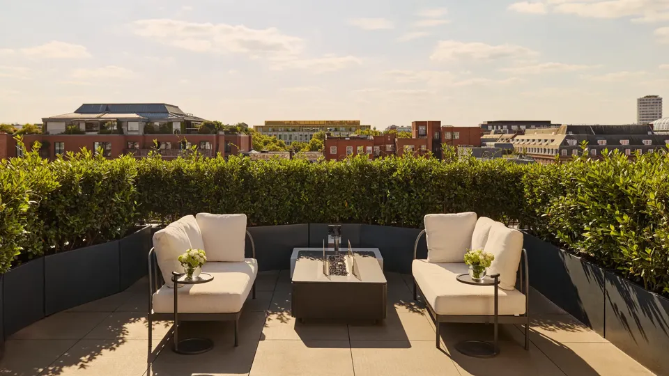Sunny rooftop terrace with two cream lounge chairs, a central fire pit-style table, and views over red-brick buildings and greenery under a blue sky.