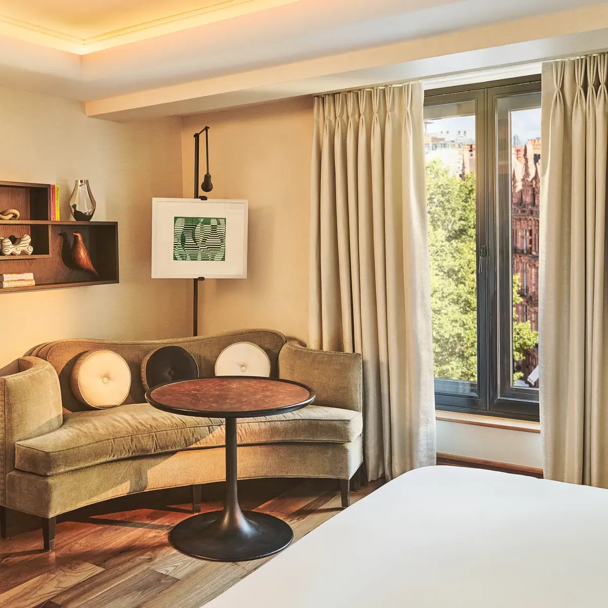 Bright room corner with a curved sofa, round table, wall art, and large window with curtains overlooking trees.