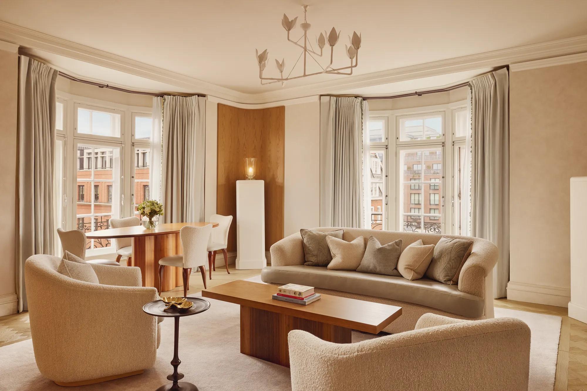 Bright and elegant living room with curved cream sofas, wooden coffee table, and dining area by tall bay windows framed with soft grey curtains overlooking red-brick buildings.