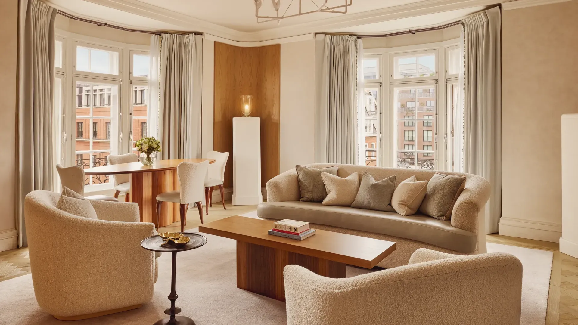 Bright and elegant living room with curved cream sofas, wooden coffee table, and dining area by tall bay windows framed with soft grey curtains overlooking red-brick buildings.
