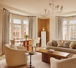 Bright and elegant living room with curved cream sofas, wooden coffee table, and dining area by tall bay windows framed with soft grey curtains overlooking red-brick buildings.