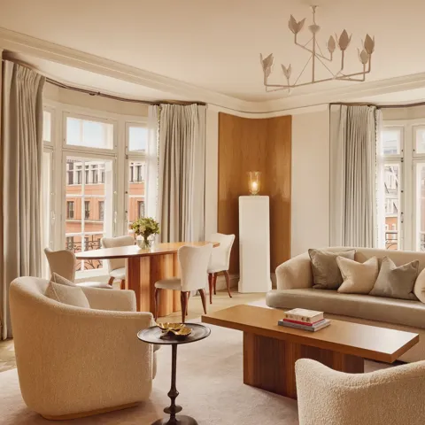 Bright and elegant living room with curved cream sofas, wooden coffee table, and dining area by tall bay windows framed with soft grey curtains overlooking red-brick buildings.